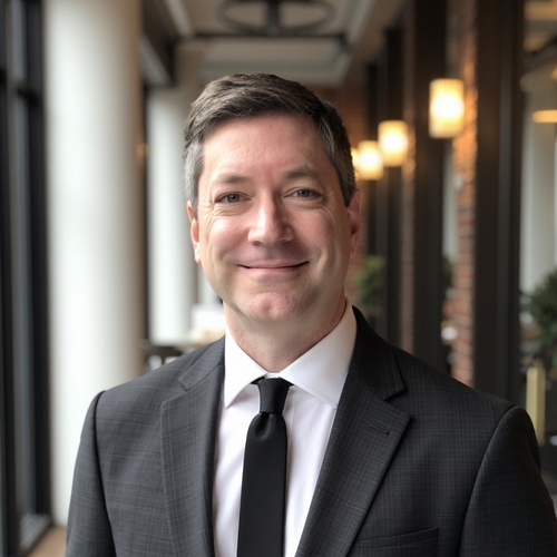 Middle-aged man in a dark suit and tie, smiling in a softly lit hallway with blurred lights in the background.