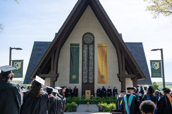 Keuka College graduates and faculty wearing regalia from behind standing in front of Norton Chapel